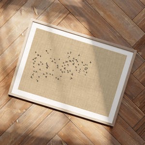 Puede incluir: Una pieza de arte mural rectangular enmarcada con un fondo de color tostado claro y textura similar al lino. Una bandada de pequeños pájaros de color gris oscuro aparece volando en el centro, dispuesta en una formación orgánica dispersa.
