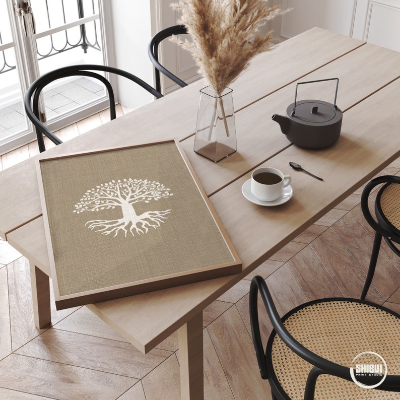 May include: A framed print of a white tree with roots on a beige linen background. The print is sitting on a wooden table with a black teapot, a cup of coffee, and a vase of dried pampas grass.