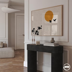 Puede incluir: Un cuadro rectangular enmarcado que presenta un gran círculo amarillo mostaza, dos pájaros negros volando y la silueta negra de una rama floreciente sobre un fondo beige claro, expuesto sobre una mesa consola negra estriada.