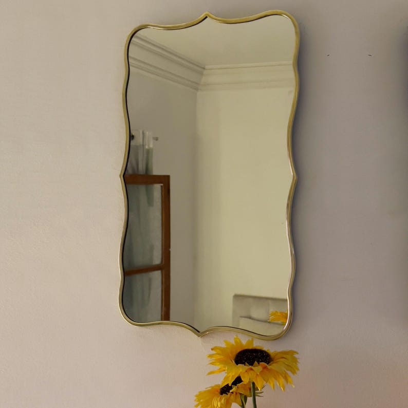 May include: A rectangular wall mirror with a gold-colored, wavy frame. The mirror reflects a room with white walls and a wooden cabinet. A bouquet of yellow sunflowers is at the bottom.