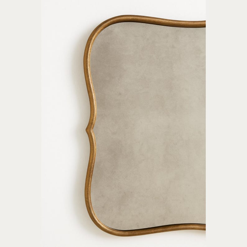 May include: A decorative mirror with a gold-colored, ornate frame. The mirror has a unique, curved shape and a neutral-toned reflective surface. The frame has a detailed, vintage-inspired design, adding a touch of elegance.