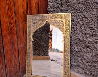 May include: A gold-coloured rectangular mirror with an arched top. The frame has intricate, ornate patterns. The mirror is leaning against a textured grey wall and a wooden door.