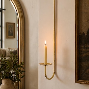 K&ouml;nnte beinhalten: Ein goldfarbener Wandkerzenhalter. Der Halter hat ein schlankes, gebogenes Design mit einer kreisf&ouml;rmigen Basis, die eine brennende, cremefarbene Kerze h&auml;lt. Der Kerzenhalter ist ein dekoratives Wohnaccessoire.