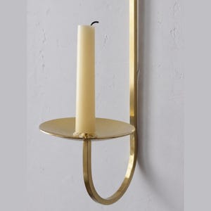 K&ouml;nnte beinhalten: Ein wandmontierter Kerzenhalter aus Messing mit einer cremefarbenen Kerze. Der Halter hat eine runde Basis und einen gebogenen Arm. Die Kerze ist unbeleuchtet und hat einen schwarzen Docht. Das Design ist minimalistisch und elegant.