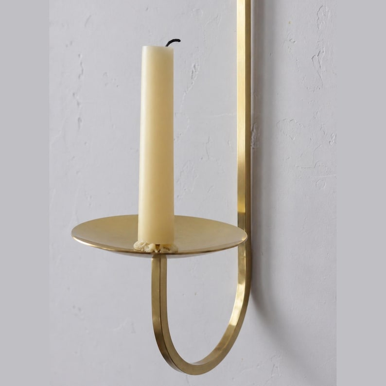 K&ouml;nnte beinhalten: Ein wandmontierter Kerzenhalter aus Messing mit einer cremefarbenen Kerze. Der Halter hat eine runde Basis und einen gebogenen Arm. Die Kerze ist unbeleuchtet und hat einen schwarzen Docht. Das Design ist minimalistisch und elegant.