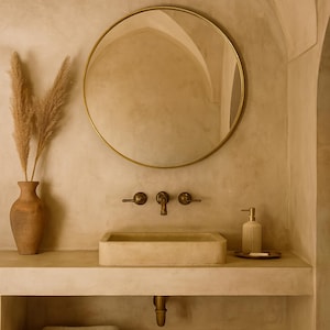Könnte beinhalten: Badezimmerinterieur mit einem runden, goldgerahmten Spiegel, einem rechteckigen Steinwaschbecken und einem Wandhahn. Eine Vase mit getrockneten Pflanzen und ein Seifenspender sind ebenfalls zu sehen. Die Farbpalette ist neutral mit warmen Tönen.