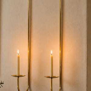 K&ouml;nnte beinhalten: Zwei Wandkerzenhalter mit brennenden Kerzen. Die Halter sind aus goldfarbenem Metall gefertigt und haben ein schlichtes, elegantes Design. Die Kerzen sind wei&szlig; und sorgen f&uuml;r ein warmes, einladendes Leuchten. Die Halter sind an einer neutralfarbenen Wand befestigt.
