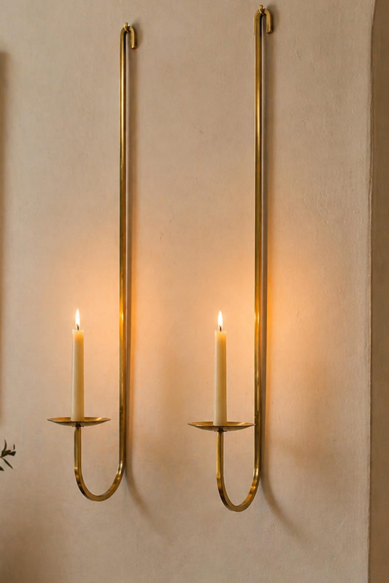 K&ouml;nnte beinhalten: Zwei Wandkerzenhalter mit brennenden Kerzen. Die Halter sind aus goldfarbenem Metall gefertigt und haben ein schlichtes, elegantes Design. Die Kerzen sind wei&szlig; und sorgen f&uuml;r ein warmes, einladendes Leuchten. Die Halter sind an einer neutralfarbenen Wand befestigt.