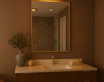 Puede incluir: Un mueble de baño con un espejo rectangular en un marco dorado. El mueble tiene un acabado de madera oscura con una encimera blanca y un grifo dorado. Un jarrón decorativo con vegetación está sobre la encimera.
