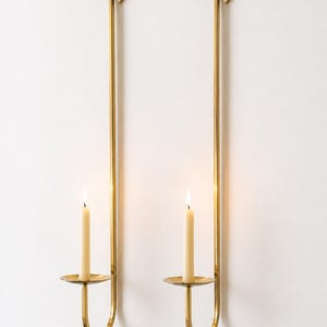 K&ouml;nnte beinhalten: Zwei Wandkerzenhalter mit brennenden Kerzen. Die Halter sind aus goldfarbenem Metall gefertigt und haben ein schlichtes, elegantes Design. Jeder Halter hat eine kreisf&ouml;rmige Basis, um die Kerze zu halten, und einen langen, geraden Stiel.