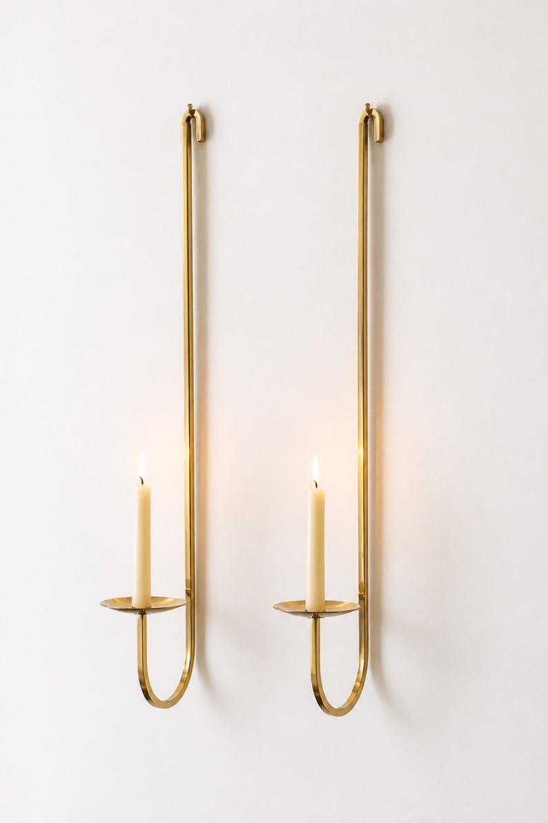 K&ouml;nnte beinhalten: Zwei Wandkerzenhalter mit brennenden Kerzen. Die Halter sind aus goldfarbenem Metall gefertigt und haben ein schlichtes, elegantes Design. Jeder Halter hat eine kreisf&ouml;rmige Basis, um die Kerze zu halten, und einen langen, geraden Stiel.