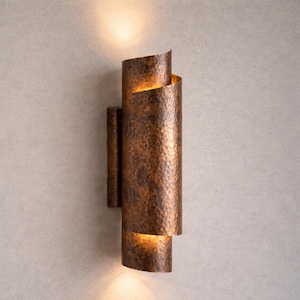 May include: A copper-colored wall sconce with a hammered texture. The sconce has a cylindrical shape with a curved top and bottom, and it emits a warm light. The fixture is mounted on a rectangular backplate.