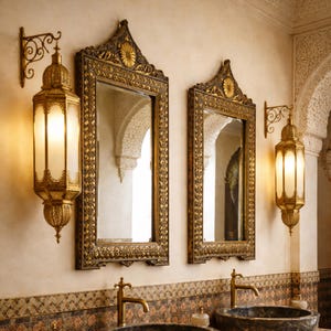 Op de afbeelding: Twee sierlijke goudomrandde spiegels en bijpassende wandkandelaars met een warme gloed. De spiegels hebben een decoratieve bovenkant en zijn boven een badkamerwastafel gemonteerd. De wandkandelaars hebben een lantaarndesign.