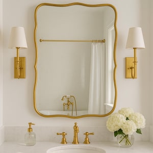May include: A gold-framed mirror with a scalloped edge hangs above a marble countertop. Two gold sconces with white shades flank the mirror. A clear glass soap dispenser, gold faucet, and a vase of white hydrangeas complete the bathroom decor.