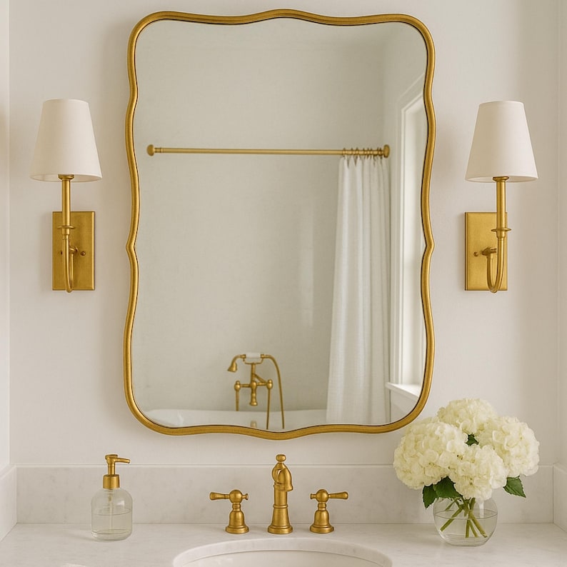 May include: A gold-framed mirror with a scalloped edge hangs above a marble countertop. Two gold sconces with white shades flank the mirror. A clear glass soap dispenser, gold faucet, and a vase of white hydrangeas complete the bathroom decor.