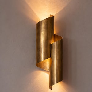 Puede incluir: Un aplique de pared que presenta dos láminas de metal curvas y superpuestas con un acabado de latón cepillado. La lámpara emite una luz cálida y difusa desde las aberturas superior e inferior, proyectando suaves reflejos en la pared.