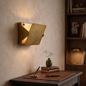 May include: A brass wall sconce with a hinged, rectangular cover, casting a warm glow. The scene includes a desk with a leather mat, books, a pen, and a small vase with flowers, creating a cozy, vintage-inspired workspace.