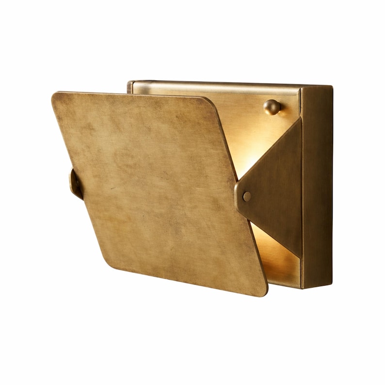 May include: A wall-mounted, brass-colored light fixture with a hinged, rectangular cover. The cover is partially open, revealing a warm, glowing light. The fixture has a minimalist design, suitable for modern interiors.