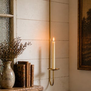 K&ouml;nnte beinhalten: Ein Wandkerzenhalter aus Messing mit einer brennenden, cremefarbenen Kerze. Der Halter ist an einer wei&szlig;en Holzwand befestigt. Eine Vase mit getrockneten Blumen, alte B&uuml;cher und ein gerahmtes Gem&auml;lde sind ebenfalls zu sehen.