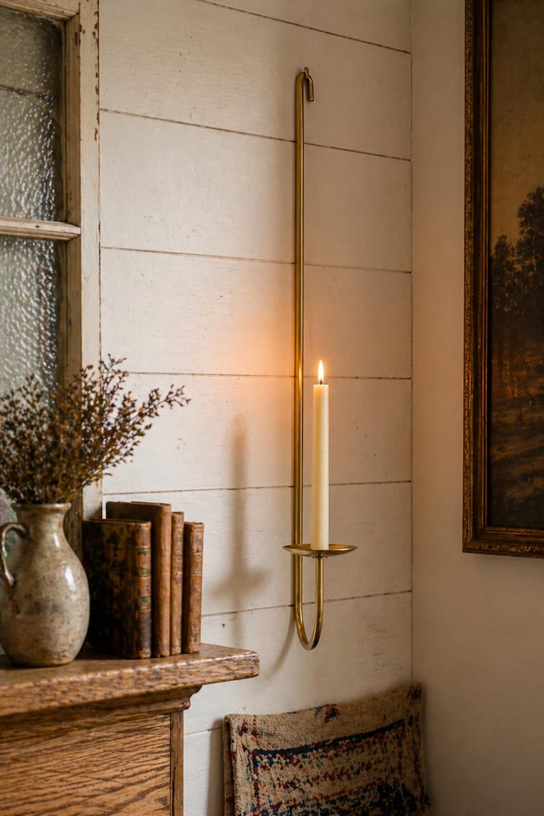 K&ouml;nnte beinhalten: Ein Wandkerzenhalter aus Messing mit einer brennenden, cremefarbenen Kerze. Der Halter ist an einer wei&szlig;en Holzwand befestigt. Eine Vase mit getrockneten Blumen, alte B&uuml;cher und ein gerahmtes Gem&auml;lde sind ebenfalls zu sehen.