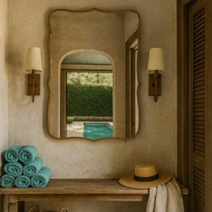 May include: A decorative wall mirror with a gold frame hangs above a wooden console. Rolled turquoise towels, a straw hat with a black band, and a cream-colored towel are on the console. Two sconces with beige shades flank the mirror.