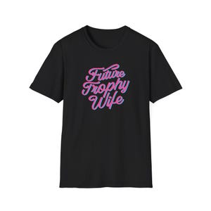 Könnte beinhalten: Ein schwarzes kurzärmeliges T-Shirt mit dem Text Future Trophy Wife, der in einer stilisierten rosa Schreibschrift mit hellblauem Umriss mittig auf der Brust aufgedruckt ist. Das Kleidungsstück hat einen Rundhalsausschnitt.