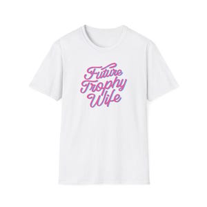 Könnte beinhalten: Weißes T-Shirt mit dem Text "Future Trophy Wife" in einer Schreibschrift. Der Text ist in rosa und blau verlaufenden Farben gehalten. Das Shirt hat einen Rundhalsausschnitt und kurze Ärmel.