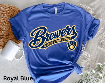 May include: A royal blue short-sleeve t-shirt featuring the text Brewers WORLD SERIES 2026 in navy blue with a yellow outline. A navy blue and yellow baseball glove icon with a white baseball is positioned below the text.