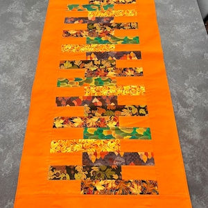 Könnte beinhalten: Ein orangefarbener Tischläufer mit einem Patchwork-Design. Der Patchwork-Stoff weist rechteckige Stoffblöcke mit Herbstblattmustern in Gelb-, Orange-, Grün- und Brauntönen auf. Der Läufer ist rechteckig und hat einen durchgehenden orangefarbenen Rand.