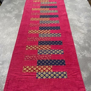 Könnte beinhalten: Ein rechteckiger magentafarbener Tischläufer mit einem zentralen Design aus abwechselnden rechteckigen Blöcken. Die Blöcke weisen goldene, marineblaue und rote Muster auf. Der Läufer ist aus Stoff und eignet sich für einen Esstisch oder eine andere Oberfläche.