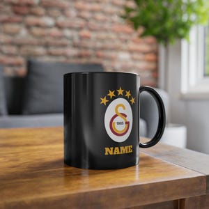 Könnte beinhalten: Schwarze Keramik-Tasse mit Henkel. Die Tasse zeigt ein Teamlogo mit einem goldenen Stern-Design und dem Text "NAME" in Gold. Das Logo enthält den Text "1905" in einem kreisförmigen Design.