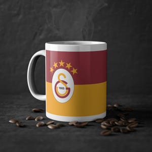 Könnte beinhalten: Eine weiße Keramik-Tasse mit einem weinroten und goldenen Design. Die Tasse zeigt das Galatasaray-Logo mit dem Text "1905" und ist mit einem heißen Getränk gefüllt, aus dem Dampf aufsteigt. Kaffeebohnen sind um die Basis der Tasse verstreut.
