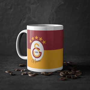 Könnte beinhalten: Eine weiße Keramiktasse mit Henkel, mit einem Design in Burgunderrot und Gold. Das Design beinhaltet ein Logo mit Sternen und den Texten "1905" und "Name". Kaffeebohnen sind um die Tasse herum verstreut.