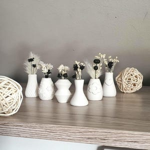 Könnte beinhalten: Ein Set aus sieben weißen Keramikvasen mit verschiedenen Formen und Designs, jede mit getrockneten weißen und schwarzen Blumen gefüllt. Die Vasen sind auf einer Holzoberfläche angeordnet.