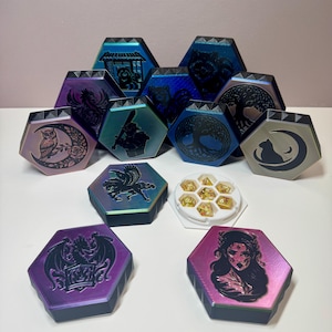 May include: A collection of iridescent hexagonal dice boxes with various engraved designs, including an owl, a tree, a cat, and a pegasus. A small white container holds several golden dice. The boxes have a black trim.