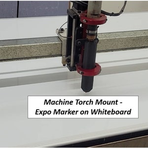 May include: A close-up of a machine torch mount with a black Expo marker attached to it. The marker is positioned over a white surface, likely a whiteboard. The text "Machine Torch Mount - Expo Marker on Whiteboard" is visible below the marker.