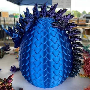 May include: A blue dragon egg sculpture with a textured, scale-like pattern. The egg is adorned with spiky, crystal-like structures in shades of blue and purple. Other dragon sculptures are visible in the background.
