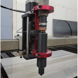 May include: A close-up of a red and black machine torch mount, with a black torch tip positioned over a metal surface. The torch is mounted on a black and silver metal frame. The surface has a design etched into it.