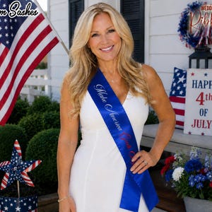 Könnte beinhalten: Eine Frau trägt eine blaue Schärpe mit dem Text "Make America Great Again" in weißer Schrift. Die Schärpe ist über einem weißen Kleid drapiert. Der Hintergrund umfasst eine amerikanische Flagge und ein Schild mit der Aufschrift "Happy 4th of July!"
