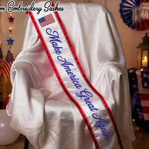 May include: A white sash with red trim and the text "Make America Great Again" in blue script. The sash features an American flag and is draped over a white chair. The words "Custom Embroidery Sashes" are at the top.