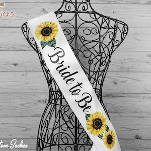 May include: White satin sash with the text "Bride to Be" in black script. The sash is decorated with yellow sunflowers.