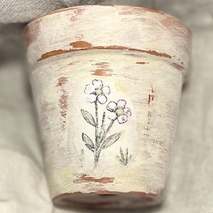 May include: A small, terracotta flower pot with a distressed, off-white painted finish. The pot features a hand-painted floral design with pink and green accents. The pot is ideal for small plants and adds a rustic touch to any space.