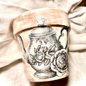 May include: A decorative terracotta flower pot with a vintage teapot and rose design. The pot features a distressed, aged appearance with a cream and peach color scheme. The teapot illustration is in black and white.