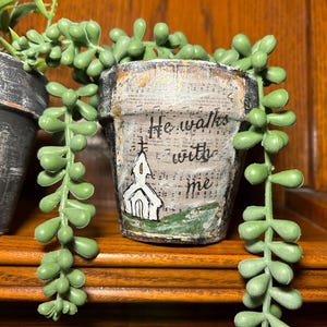 May include: A small white ceramic planter with a green base and a white church illustration. The planter is decorated with a vintage sheet music background and the text "He walks with me". The planter is filled with a green succulent plant.
