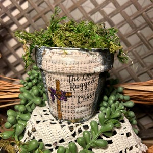 May include: A small terracotta pot with a green moss plant inside. The pot is decorated with a vintage-style print of a cross and the words "The Old Rugged Cross".