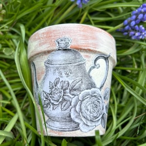 May include: A small, decorative terracotta flower pot with a distressed finish. The pot features a black and white illustration of a teapot and roses. The pot is surrounded by green grass and purple flowers.