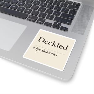 Könnte beinhalten: Ein quadratischer Aufkleber mit cremefarbenem Hintergrund und weißem Rand, der den schwarzen Text "Deckled" in einer großen Serifenschrift über dem kleineren schwarzen Text "edge defender" mit einer dünnen Trennlinie zeigt.