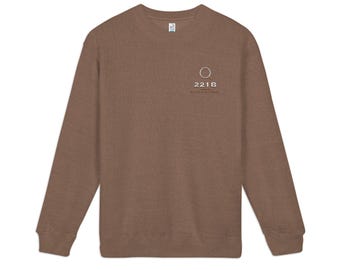 May include: A brown long-sleeve crewneck sweatshirt featuring a small white graphic on the left chest. The graphic includes a magnifying glass icon above the text 221B and the word Baker Street in a smaller font below the numbers.
