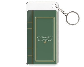 May include: A rectangular acrylic keyring featuring a green book cover design with gold-coloured border lines and the text COLD HANDS LONG BOOK printed in white, accompanied by a small star icon and a silver-toned metal split ring attachment.