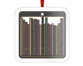 May include: A square, clear acrylic ornament featuring a graphic illustration of a bookshelf filled with vertical books in shades of green, brown, purple, and black. The ornament is suspended by a thin red ribbon attached to a small hole at the top.